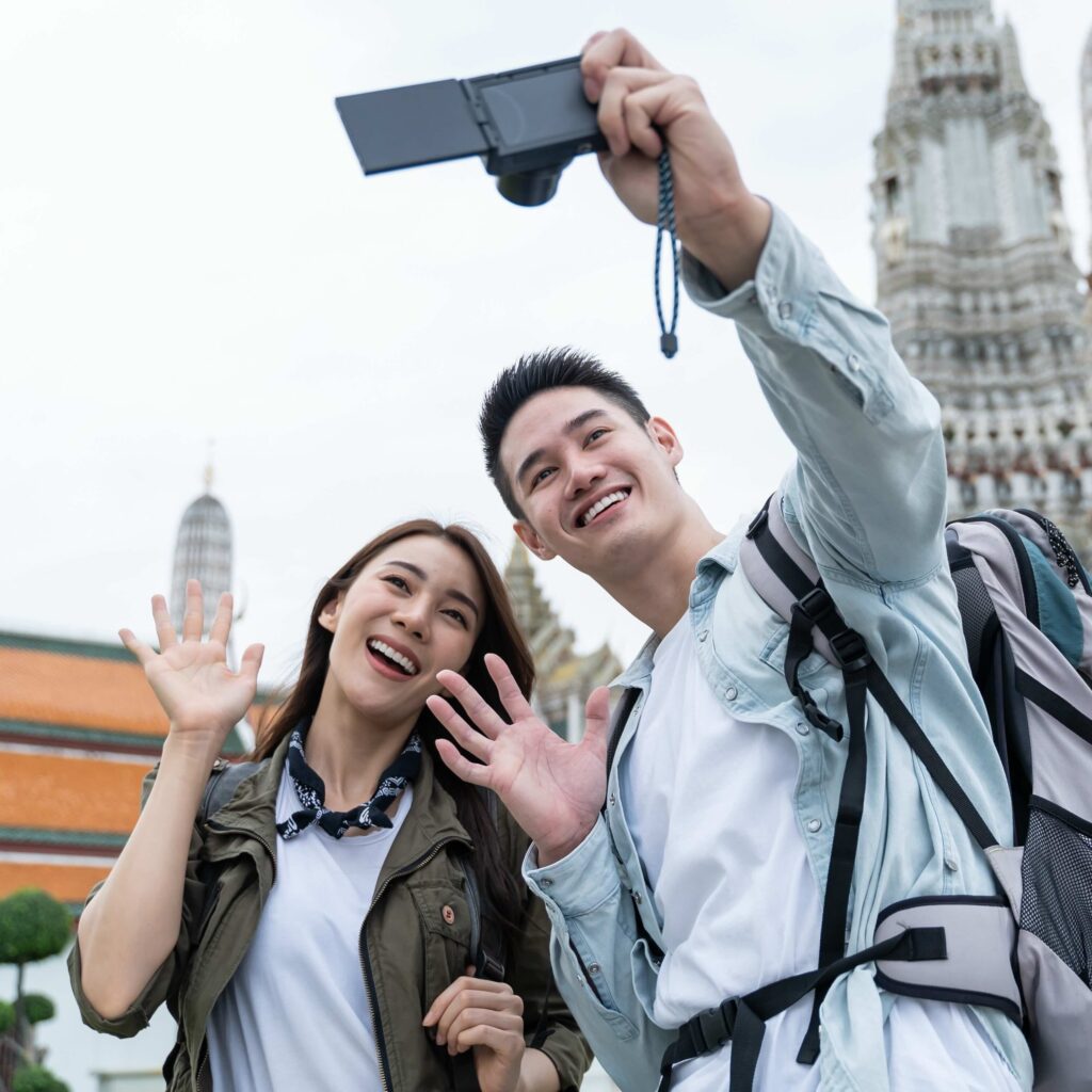 Asian couple backpacker using camera selfie and take a picture in city. Attractive man and woman tourist traveler travel on street, using smartphone record vlog on holiday vacation trip in Thailand.